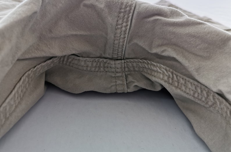 May include: Close-up of the crotch area of khaki-colored pants. The image highlights the seams and stitching details, showcasing the construction of the garment. The fabric appears to be a durable cotton blend.