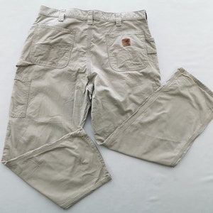 May include: Khaki-colored work pants with multiple pockets, including a side pocket and two back pockets. The pants feature a brown leather patch with a logo. The pants are made of a durable material and are designed for work.