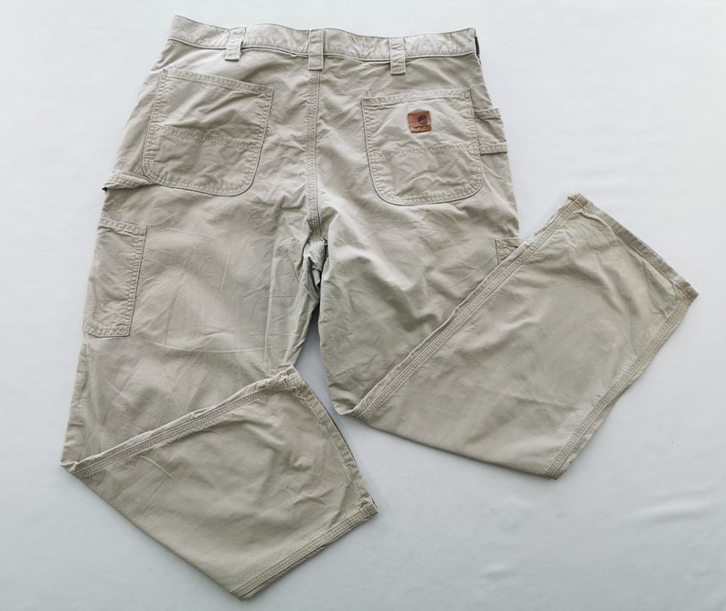 May include: Khaki-colored work pants with multiple pockets, including a side pocket and two back pockets. The pants feature a brown leather patch with a logo. The pants are made of a durable material and are designed for work.
