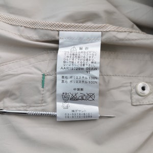 May include: A beige garment interior featuring a white care label with Japanese text, including the code AAR19729W 09AW YI, 100% polyester composition, and laundry symbols. A thin metal tool is positioned horizontally across the label for scale.