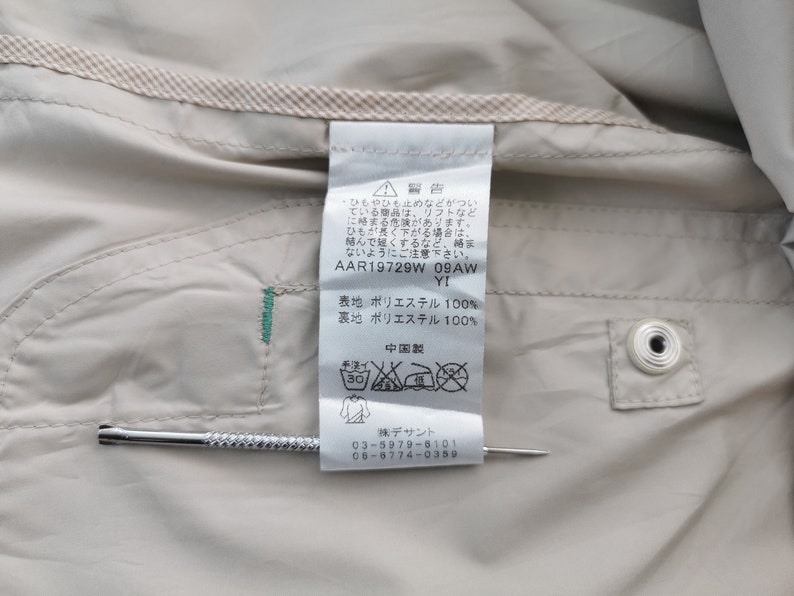 May include: A beige garment interior featuring a white care label with Japanese text, including the code AAR19729W 09AW YI, 100% polyester composition, and laundry symbols. A thin metal tool is positioned horizontally across the label for scale.