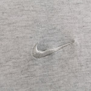 May include: Close-up of a heather gray fabric with a white embroidered Nike swoosh logo. The logo is positioned in the center of the image. The fabric has a textured appearance.