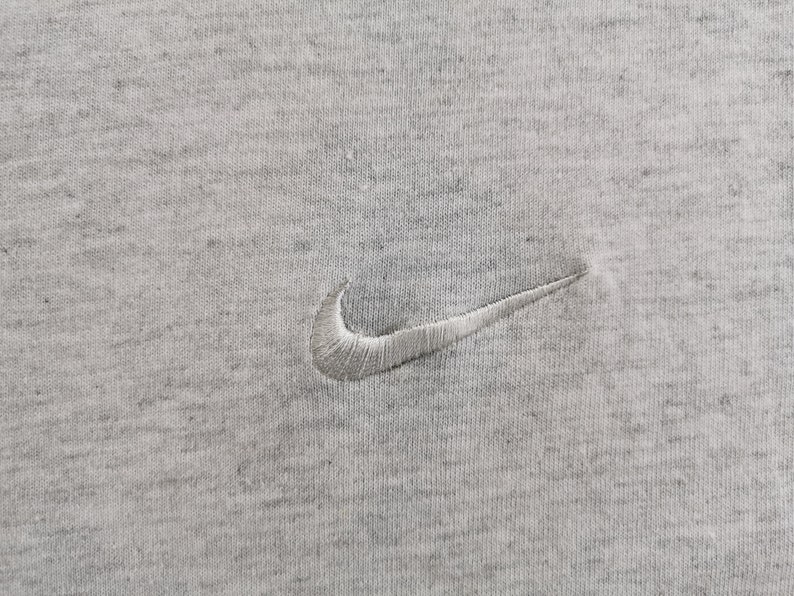 May include: Close-up of a heather gray fabric with a white embroidered Nike swoosh logo. The logo is positioned in the center of the image. The fabric has a textured appearance.