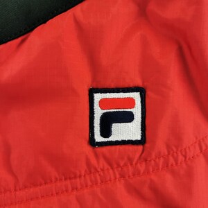 May include: Close-up of a red and black jacket. The jacket features a square logo with the brand's name in white, red, and navy blue. The jacket is made of a lightweight, possibly nylon material.