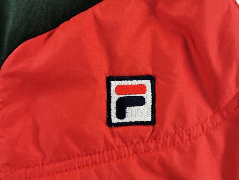 May include: Close-up of a red and black jacket. The jacket features a square logo with the brand's name in white, red, and navy blue. The jacket is made of a lightweight, possibly nylon material.