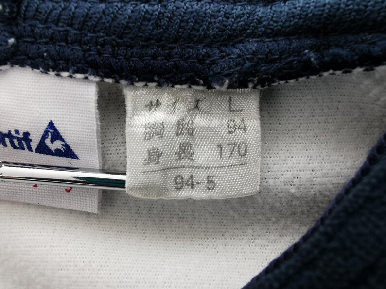 K&ouml;nnte beinhalten: Nahaufnahme eines wei&szlig;en Stoffetiketts mit japanischem Text und Gr&ouml;&szlig;enangaben. Das Etikett ist an einem wei&szlig;en Kleidungsst&uuml;ck mit marineblauem Kragen befestigt. Das Etikett enth&auml;lt die Gr&ouml;&szlig;e "L", Brustumfang "94" und H&ouml;he "170" cm.