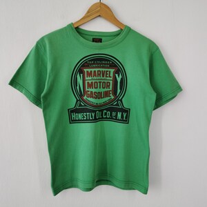 May include: A bright green short-sleeve t-shirt featuring a large black and red graphic on the chest. The design includes the text Marvel Motor Gasoline, Top Cylinder Lubrication, and Honestly Oil Co. of N.Y. inside a circular emblem.