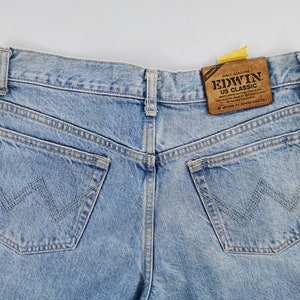 May include: Light blue denim jeans with classic five-pocket styling. Features a brown leather patch with "EDWIN US CLASSIC" text. The back pockets have a distinctive W-shaped stitch design. A red Edwin label is visible.