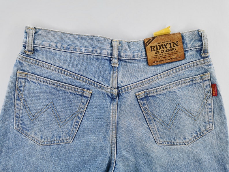 May include: Light blue denim jeans with classic five-pocket styling. Features a brown leather patch with "EDWIN US CLASSIC" text. The back pockets have a distinctive W-shaped stitch design. A red Edwin label is visible.