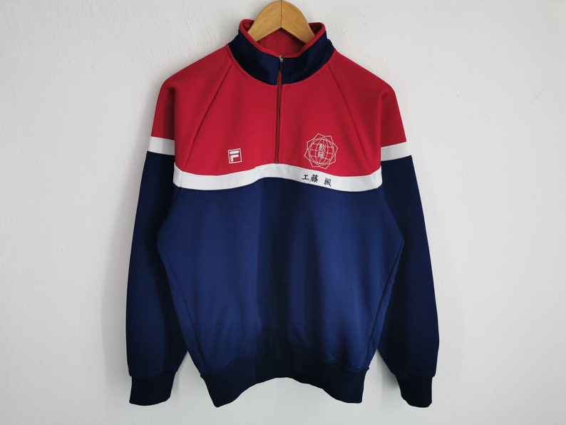 Puede incluir: Una chaqueta tipo su&eacute;ter de color rojo y azul marino con cremallera de un cuarto y una franja horizontal blanca en el pecho. La prenda presenta un logotipo Fila blanco en el lado derecho y un emblema circular blanco con texto japon&eacute;s.
