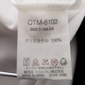K&ouml;nnte beinhalten: Nahaufnahme eines wei&szlig;en Stoffetiketts mit schwarzem Text und Symbolen. Das Etikett zeigt den Text "QTM-6102", "95S106-5A" und "ポリエステル 100%", zusammen mit Pflegehinweisen und "MADE IN JAPAN".