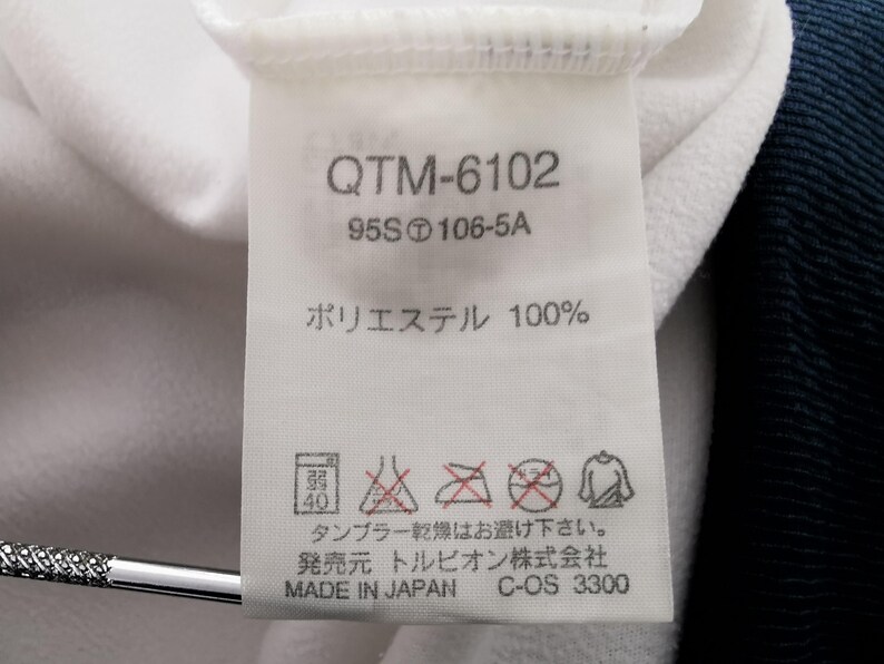 K&ouml;nnte beinhalten: Nahaufnahme eines wei&szlig;en Stoffetiketts mit schwarzem Text und Symbolen. Das Etikett zeigt den Text "QTM-6102", "95S106-5A" und "ポリエステル 100%", zusammen mit Pflegehinweisen und "MADE IN JAPAN".