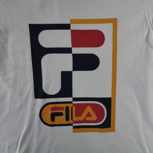 May include: White t-shirt featuring a large graphic design. The design is split vertically, with a navy blue and white checkered pattern on the left and a red and white design on the right. The word "FILA" is at the bottom.