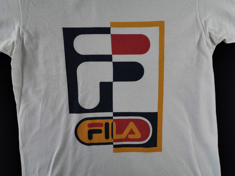 May include: White t-shirt featuring a large graphic design. The design is split vertically, with a navy blue and white checkered pattern on the left and a red and white design on the right. The word "FILA" is at the bottom.