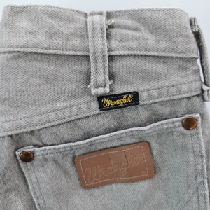 May include: Close-up of gray Wrangler jeans. The jeans feature a classic five-pocket design, belt loops, and a leather patch with the Wrangler logo. The back pockets have copper-colored rivets. The Wrangler label is black with gold lettering.