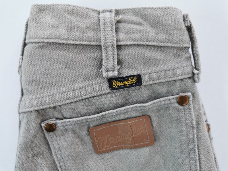 May include: Close-up of gray Wrangler jeans. The jeans feature a classic five-pocket design, belt loops, and a leather patch with the Wrangler logo. The back pockets have copper-colored rivets. The Wrangler label is black with gold lettering.