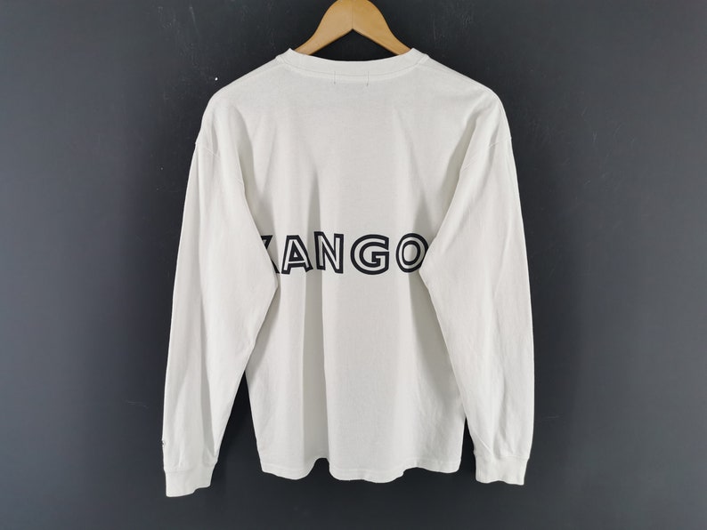 Peut inclure: T-shirt blanc &agrave; manches longues avec le mot "KANGO" imprim&eacute; en noir dans le dos. Le t-shirt est suspendu &agrave; un cintre en bois sur un fond gris fonc&eacute;. Les manches sont longues et les poignets sont unis.