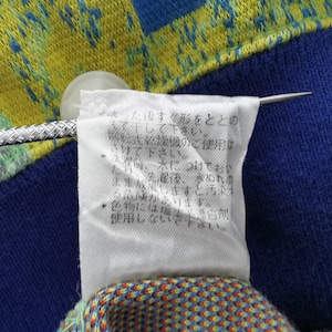 May include: Close-up of a fabric with a blue, yellow, and orange pattern. A white tag with Japanese text is attached with a silver pin. The tag provides care instructions.