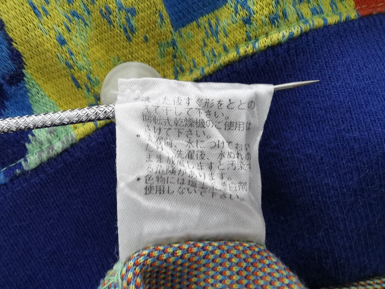 May include: Close-up of a fabric with a blue, yellow, and orange pattern. A white tag with Japanese text is attached with a silver pin. The tag provides care instructions.