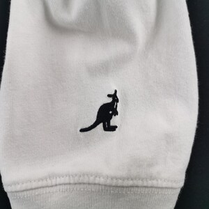 Peut inclure: Gros plan sur un tissu blanc avec un logo de kangourou noir brod&eacute; sur le bas. Le kangourou est repr&eacute;sent&eacute; de profil. Le tissu pr&eacute;sente une texture c&ocirc;tel&eacute;e et une couture.