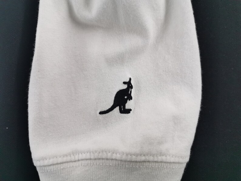 Peut inclure: Gros plan sur un tissu blanc avec un logo de kangourou noir brod&eacute; sur le bas. Le kangourou est repr&eacute;sent&eacute; de profil. Le tissu pr&eacute;sente une texture c&ocirc;tel&eacute;e et une couture.