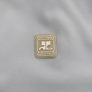 May include: A light blue fabric with a rectangular patch in the center. The patch is beige with the words "courr&egrave;ges" and "sport futur" in a dark font. The patch has a stylized logo in the center.