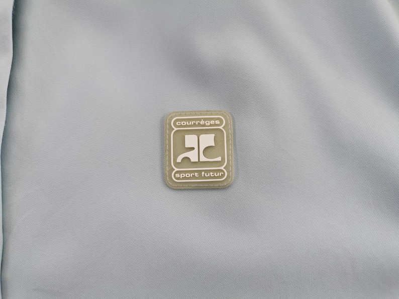 May include: A light blue fabric with a rectangular patch in the center. The patch is beige with the words "courr&egrave;ges" and "sport futur" in a dark font. The patch has a stylized logo in the center.