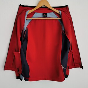 Op de afbeelding: Rood en marineblauw sportjack met een volledige ritssluiting, zwarte bies en een lichtblauwe binnenvoering. De jas hangt aan een houten hanger. Het Nike-logo is zichtbaar.