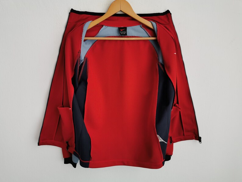 Op de afbeelding: Rood en marineblauw sportjack met een volledige ritssluiting, zwarte bies en een lichtblauwe binnenvoering. De jas hangt aan een houten hanger. Het Nike-logo is zichtbaar.
