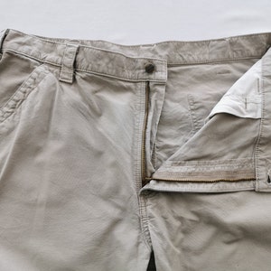 May include: Close-up of khaki-colored work pants with an open zipper and button closure. The pants have multiple pockets and belt loops. The fabric appears to be a durable cotton blend, suitable for work or casual wear.