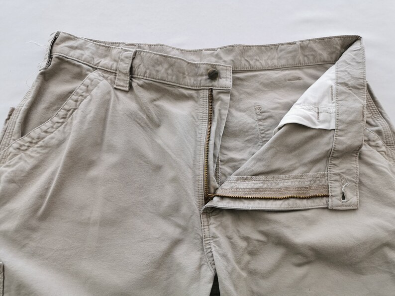 May include: Close-up of khaki-colored work pants with an open zipper and button closure. The pants have multiple pockets and belt loops. The fabric appears to be a durable cotton blend, suitable for work or casual wear.