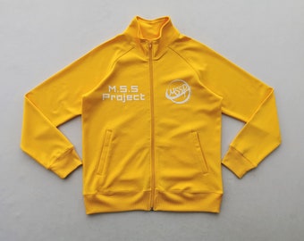 Puede incluir: Una chaqueta deportiva amarilla con cremallera, cuello alto, mangas largas con puños acanalados y dos bolsillos delanteros. El pecho muestra el texto blanco M.S.S Project y un logotipo circular blanco con las letras MSSP.