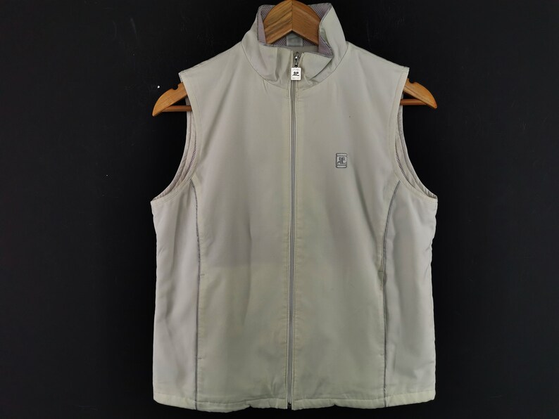 Puede incluir: Un chaleco sin mangas de color beige claro con cremallera completa y cuello alto. El chaleco tiene un peque&ntilde;o logotipo en el pecho y est&aacute; colgado de una percha de madera. El fondo es negro s&oacute;lido.