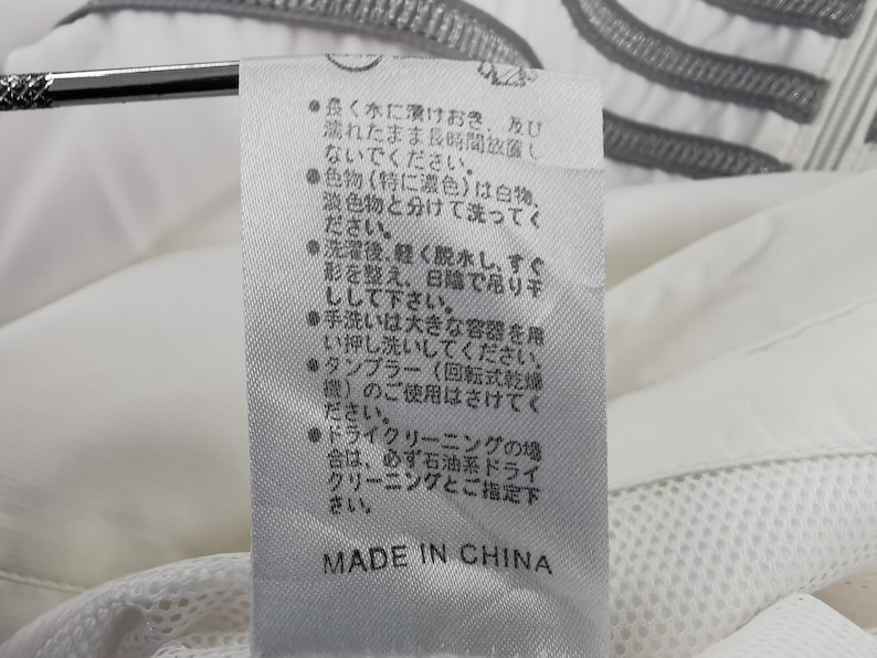 Puede incluir: Una etiqueta de tela blanca con texto japon&eacute;s y las palabras "MADE IN CHINA" en negro. La etiqueta est&aacute; adherida a una prenda blanca con una textura de malla.