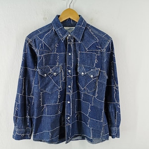 May include: A long-sleeve denim shirt in a dark blue wash featuring a white script pattern throughout. The shirt includes a pointed collar, a front button-down closure with silver-tone snaps, and two chest pockets with pointed flaps and snap closures.