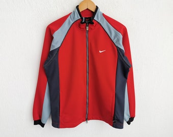 Puede incluir: Una chaqueta deportiva roja con cremallera, paneles gris claro en hombros y mangas, paneles laterales azul marino oscuro, cuello alto y un logotipo Nike blanco en el pecho izquierdo. La chaqueta incluye una cremallera frontal completa.