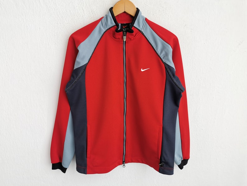 Op de afbeelding: Rood, grijs en marineblauw Nike-trainingsjack met een volledige ritssluiting. De jas heeft een klein wit Nike-logo op de borst en hangt aan een houten hanger. Zwarte kraag en manchetten.