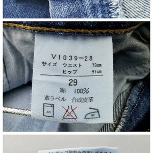 May include: Close-up of blue denim jeans with a button closure. The tag reads "V1039-28" with waist 73cm and hip 91cm. The jeans are size 29, made of 100% cotton. Additional text on the tag includes washing instructions and the manufacturer's information.