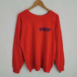 May include: A bright red long-sleeve sweatshirt featuring a crew neckline and ribbed cuffs and hem. On the upper left chest, there is a blue graphic logo with a grid-patterned square icon and the text "BUILDERS SQUARE" printed in blue.