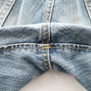 May include: Close-up of light blue denim jeans. The image highlights the stitching details, with yellow thread used for the seams. The fabric has a textured appearance, and the focus is on the waist area of the jeans.