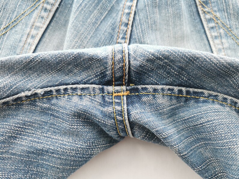 May include: Close-up of light blue denim jeans. The image highlights the stitching details, with yellow thread used for the seams. The fabric has a textured appearance, and the focus is on the waist area of the jeans.