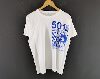 Puede incluir: Una camiseta blanca de manga corta con un estampado gráfico azul en el lado derecho. El diseño incluye el texto 501st JOINT FIGHTER WING STRIKE WITCHES, un emblema circular y una ilustración del personaje Yoshika Miyafuji.
