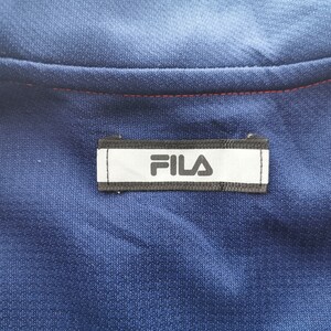 Puede incluir: Una prenda deportiva de color azul marino que presenta una etiqueta rectangular blanca con el texto negro FILA centrado en la zona del cuello. La prenda incluye detalles en rosa brillante a lo largo de las costuras de los hombros y una costura roja.