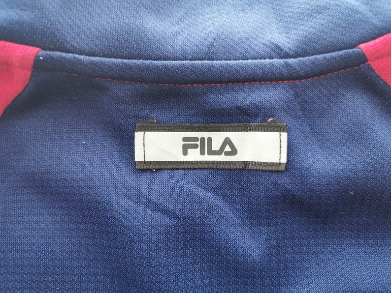 Puede incluir: Una prenda deportiva de color azul marino que presenta una etiqueta rectangular blanca con el texto negro FILA centrado en la zona del cuello. La prenda incluye detalles en rosa brillante a lo largo de las costuras de los hombros y una costura roja.