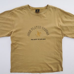May include: Mustard-colored t-shirt with short sleeves. The shirt features embroidered text that reads "JOHN PLAYER SPECIAL" in an arch, with "THE SPIRIT OF H.M.S HERO" below. A logo is embroidered in the center.