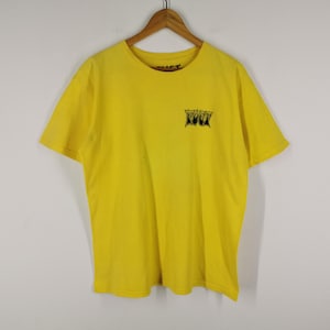 May include: Bright yellow t-shirt with a crew neck and short sleeves. The shirt features a small black graphic on the upper left chest. The shirt is hanging on a wooden hanger against a white background.