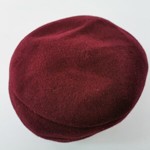 May include: A burgundy newsboy cap made of a soft, textured material. The hat has a rounded crown and a small, stiff brim. The cap is a solid color and appears to be made of wool or a similar fabric. This hat is a classic accessory.