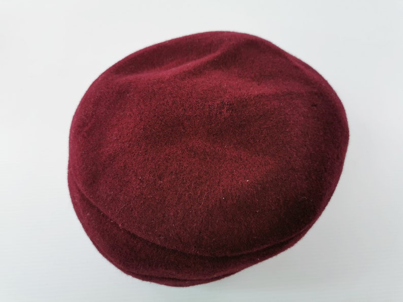 May include: A burgundy newsboy cap made of a soft, textured material. The hat has a rounded crown and a small, stiff brim. The cap is a solid color and appears to be made of wool or a similar fabric. This hat is a classic accessory.