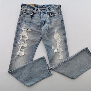May include: Light blue denim jeans with distressed detailing, featuring ripped sections on the legs. The jeans have a classic five-pocket design and a button-fly closure. The denim has a faded wash, giving it a worn appearance.