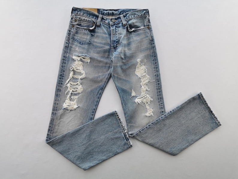 May include: Light blue denim jeans with distressed detailing, featuring ripped sections on the legs. The jeans have a classic five-pocket design and a button-fly closure. The denim has a faded wash, giving it a worn appearance.